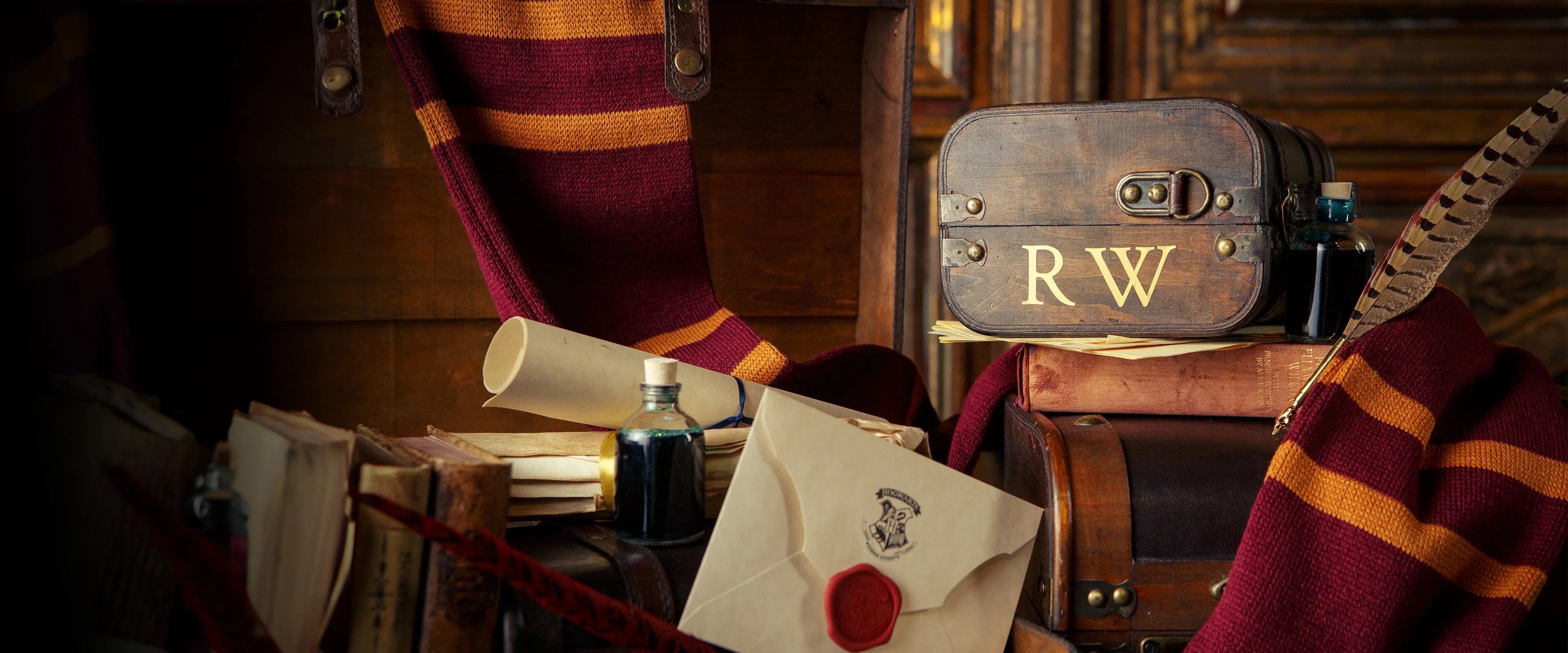 A Hogwarts gift trunk sits on a rustic table top surrounded by Hogwarts acceptance letters, scrolls, ink pots and Gryffindor knit scarves