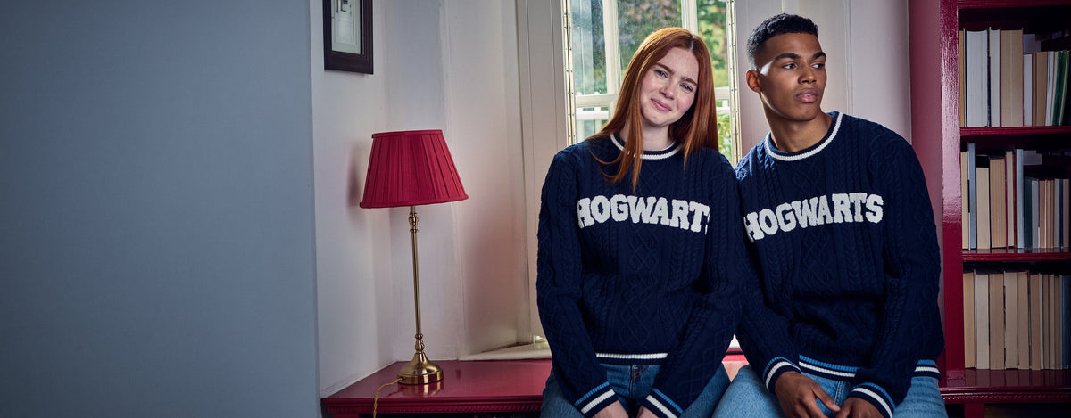 A young woman with red hair and a young man sitting side-by-side wearing matching navy blue Hogwarts sweaters in a cozy room setting.