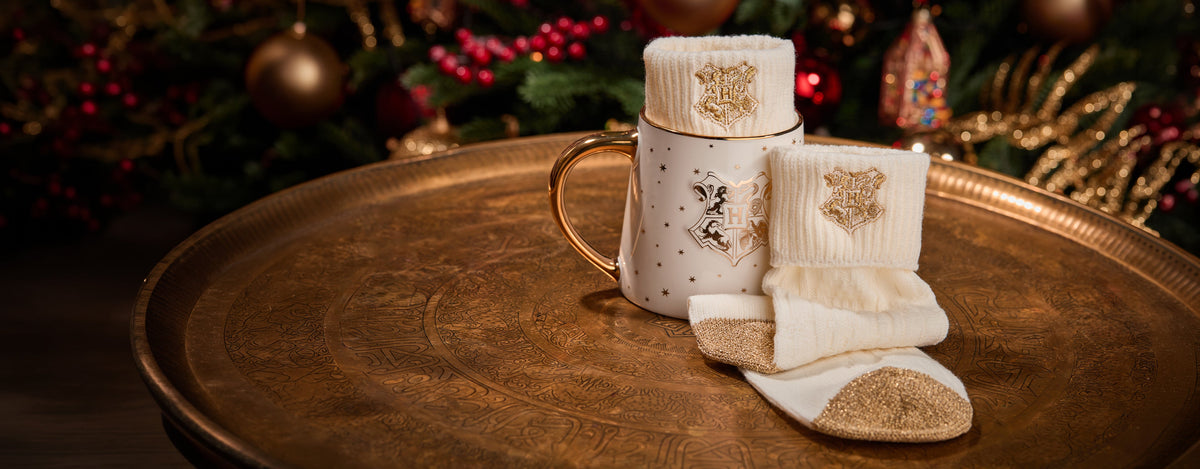 A festive Christmas-themed Hogwarts set featuring a white ceramic mug with a gold handle and matching white knit socks, all decorated with gold glitter and embroidered gold motifs, arranged on a large round bronze tray with a blurred Christmas background.