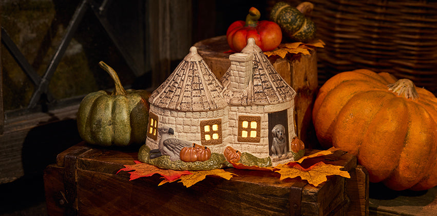 A decorative ceramic Hagrid's Hut Tealight displayed on a rustic wooden surface with pumpkins and autumn leaves around it.