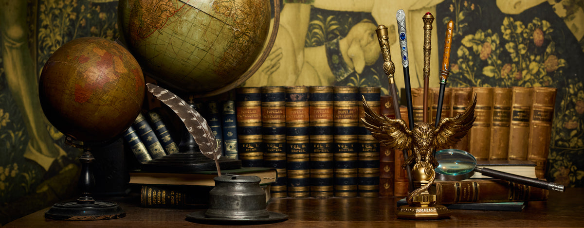 A vintage study still life featuring two antique globes, leather-bound books, a feather quill in a metal ink bottle, a gold owl wand stand, and a magnifying glass on a wooden surface against a patterned tapestry.