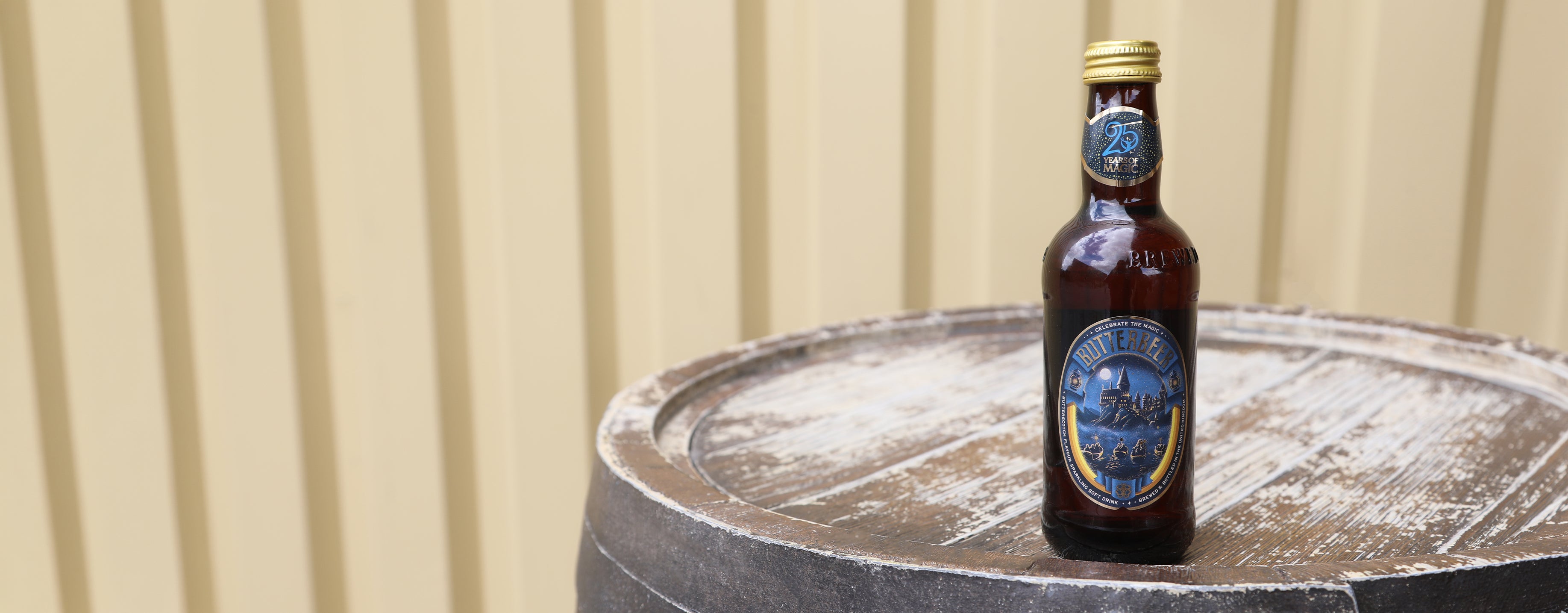 Butterbeer bottle with a blue label on a wooden barrel against a beige wall.