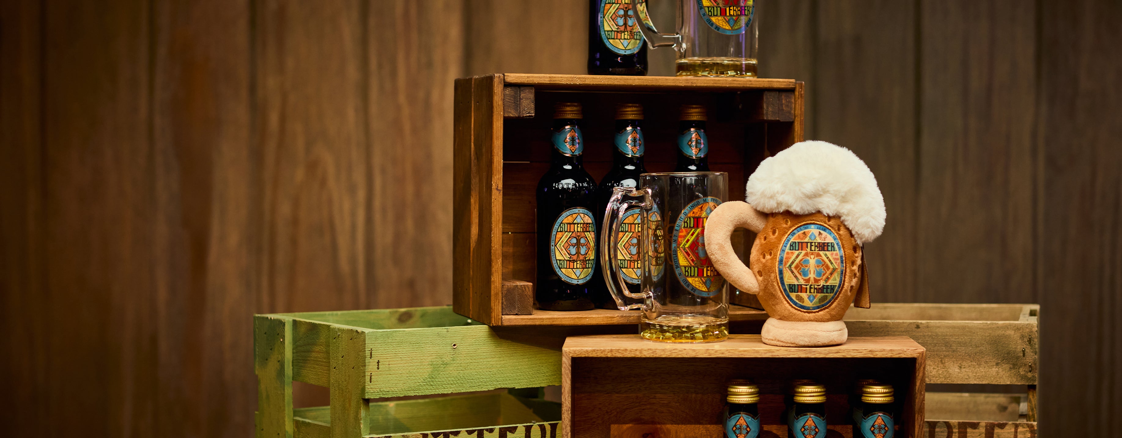 A collection of traditional Butterbeerware displayed on rustic wooden crates against a dark wood panel background. The set includes dark glass bottles with colorful labels, a clear glass mug, and a plush Butterbeer stein decorated with a white foam head and a central MinaLima label.