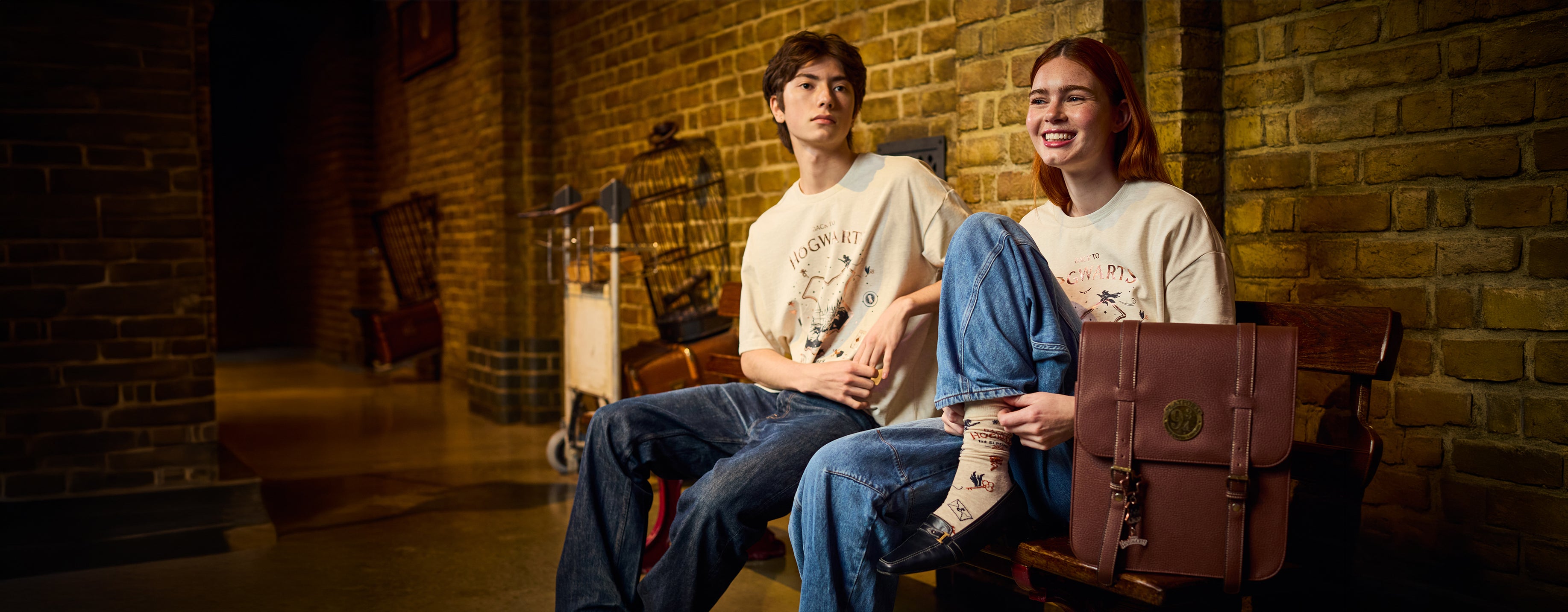 Two young adults sitting on a bench in a dimly lit brick interior, wearing matching cream Back to Hogwarts T-shirts and blue jeans, with a brown leather suitcase beside them.