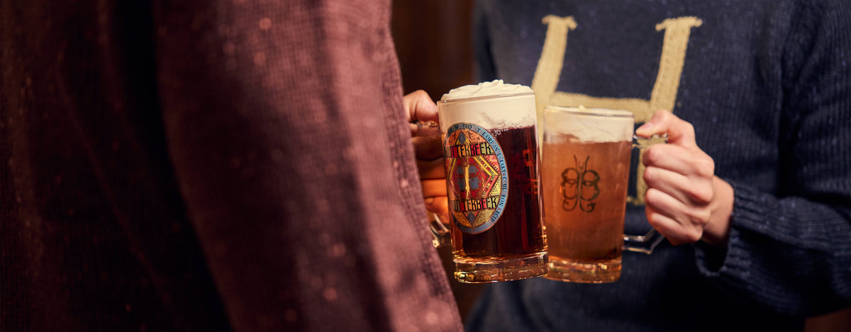 Two people holding Butterbeer mugs in a dimly lit indoor setting. One mug has a dark Butterbeer with a foam head and a circular logo, and the other has a lighter Butterbeer with a foam head and a stylized 'BB' logo.