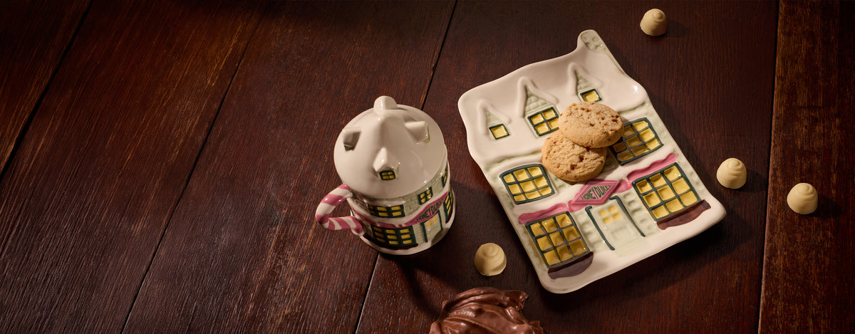 A ceramic mug shaped like Honeydukes with a pink and white handle and yellow window details sits beside a matching house-shaped plate holding two cookies, on a dark wooden tabletop with a few chocolates scattered around.