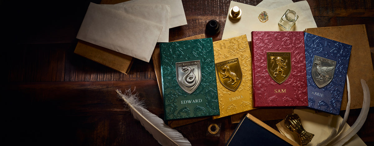 Four Hogwarts-style crested notebooks in green, yellow, red, and blue are arranged on a dark wooden desk with old papers, a quill, ink, and small props.