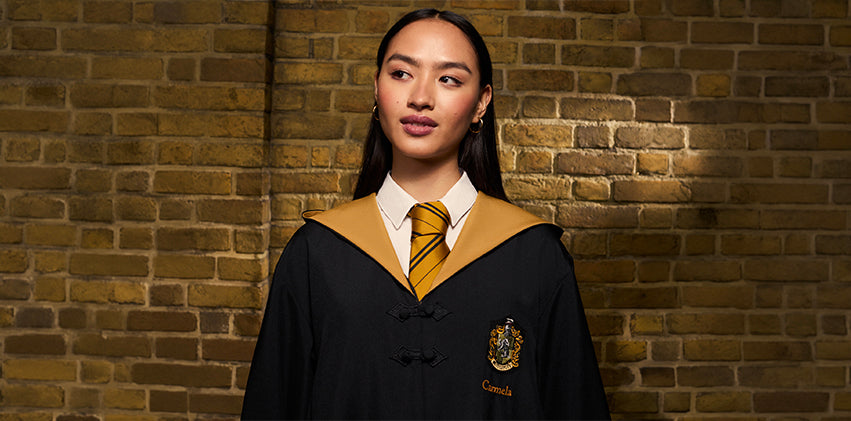 A woman wearing a black Hogwarts-style gown with a gold collar and a yellow striped tie, standing against a brick wall. The gown features a Hufflepuff crest on the left chest and her name embroidered below it.