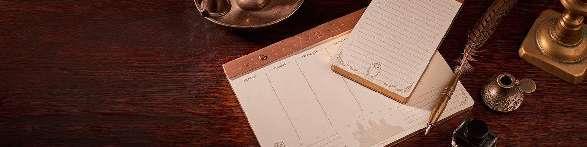 A styled flat-lay of a vintage desk scene featuring a wooden desk pad with lined notepad paper, a gold pen, a feather quill, an ink bottle, and brass desk accessories on a dark wood background.