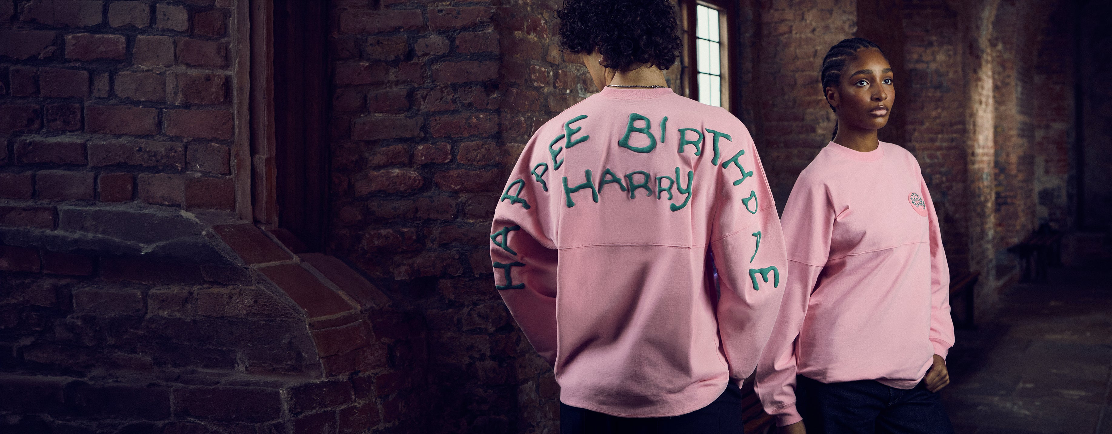 Two people wearing pink sweatshirts with Happy Birthday text in a brick-walled corridor.