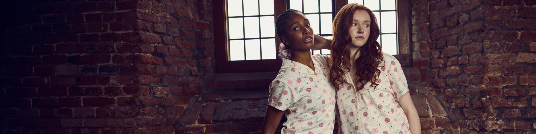 Two women in pajama sets standing in a room with brick walls and a window.