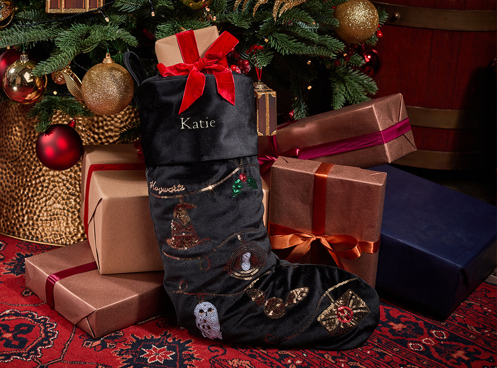 A dark velvet Christmas stocking with embroidered holiday motifs and the name 'Katie' on the cuff, styled among wrapped presents under a decorated Christmas tree.