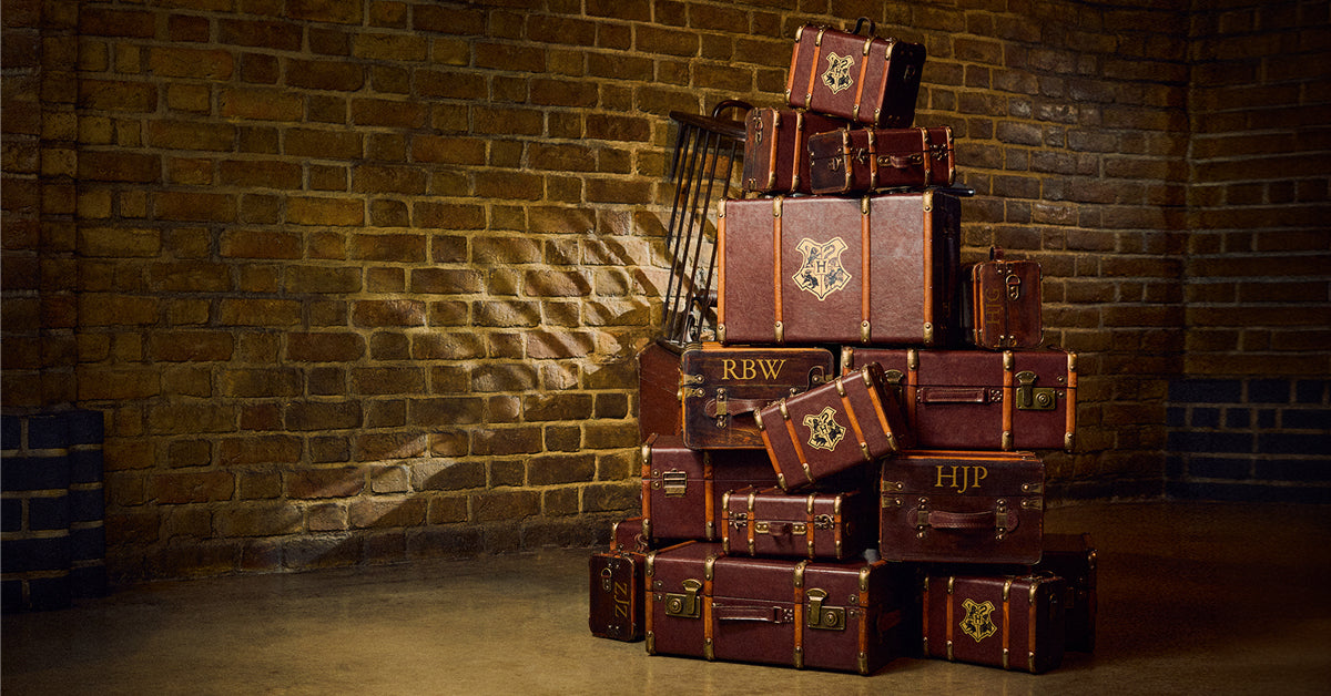 A stack of vintage-style brown leather trunks and suitcases with gold trim and crest emblems, arranged against a brick wall in a dimly lit room.